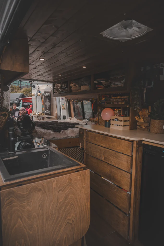 A campervan kitchen beneath a dark wooden ceiling, with a black sink, wooden drawers and a bed at the back.