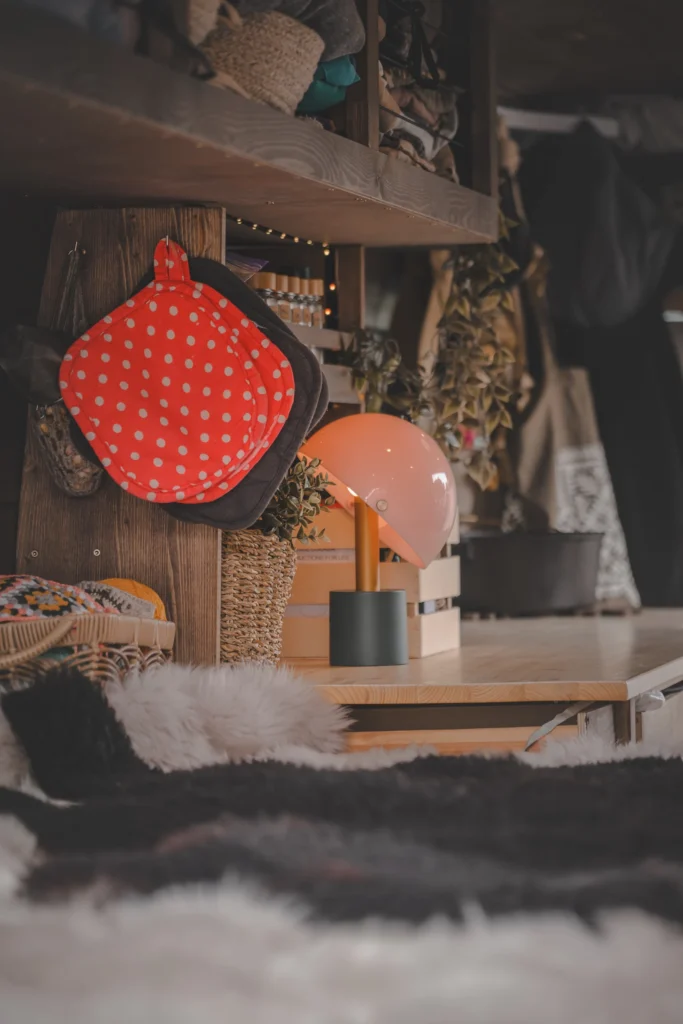 A pink table lamp and a red polka-dot potholder in a wooden campervan kitchen, with fluffy bed covers in the foreground.