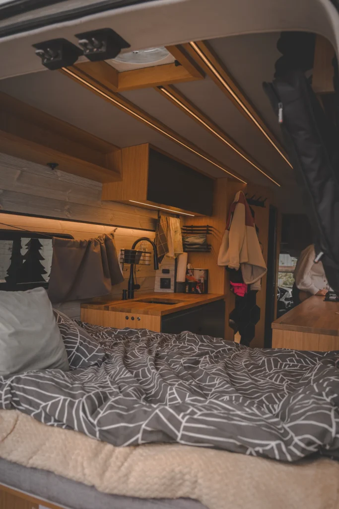 A bed with grey and white bedding, a wooden kitchenette and a skylight inside a warmly lit campervan.