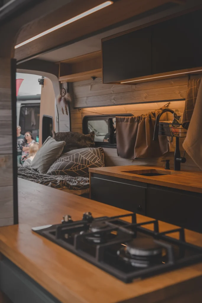 A two-burner gas hob on a wooden worktop, with a sink, dark cabinets and a bed behind it inside a campervan.