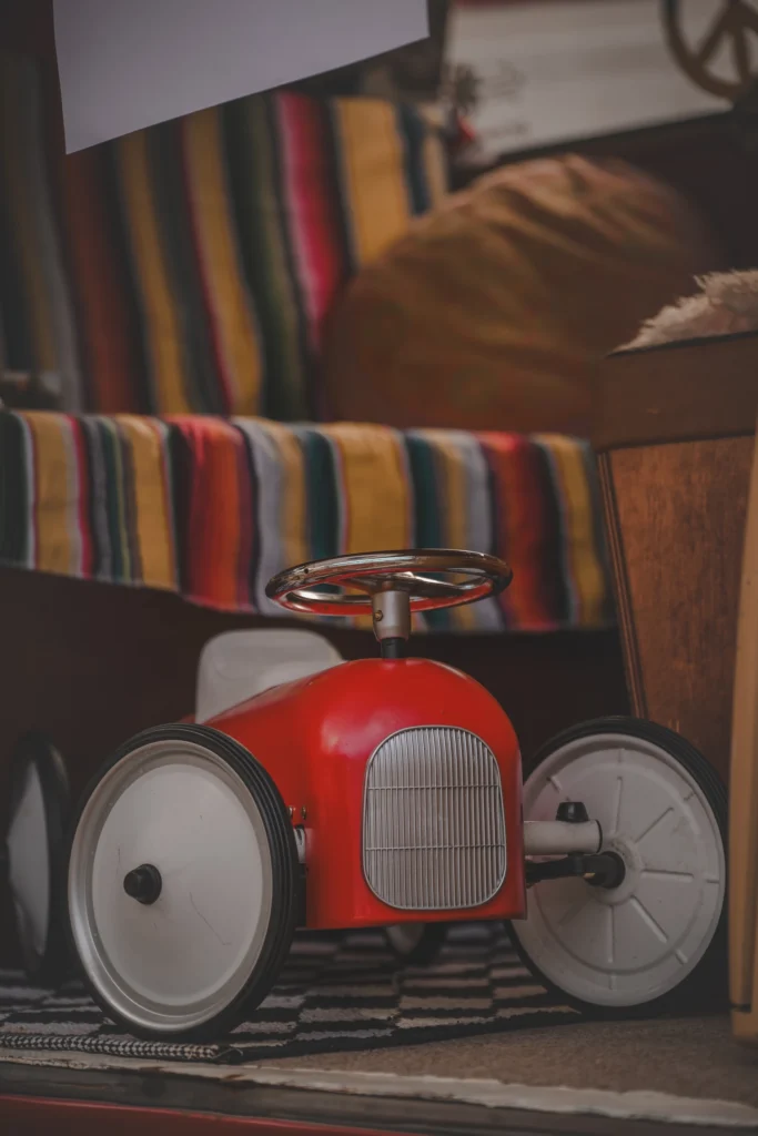 A red vintage-style ride-on toy car on the floor of a campervan with a striped seat cover.