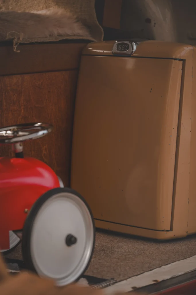 A small mustard-brown fridge on a campervan floor beside the large white wheel of a red toy car.