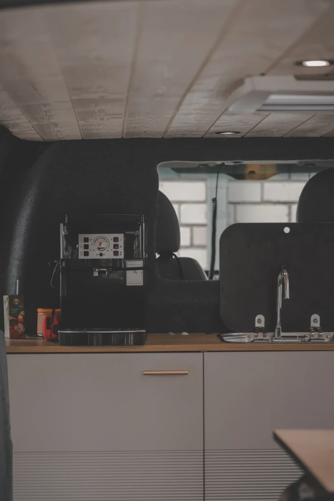 A campervan kitchenette with light cabinets, a coffee machine and a small sink on a wooden worktop.