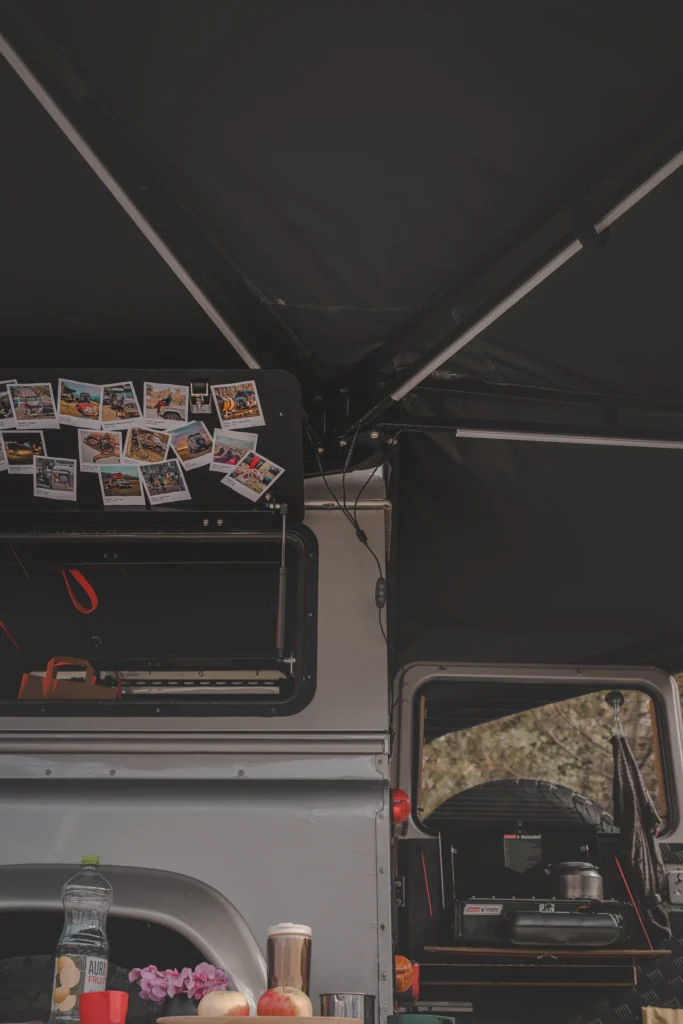 A panel covered with travel photos and a pull-out kitchen unit on a vehicle beneath a dark awning.