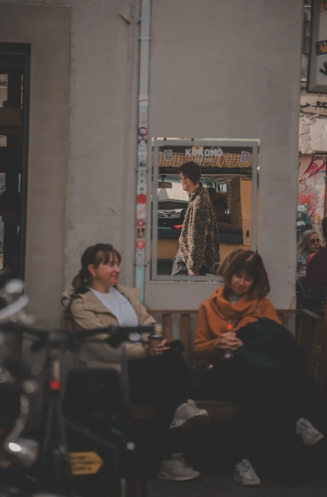 Two women sit on a bench in the Pohjala Factory courtyard, with a passer-by reflected in a wall-mounted mirror behind them.