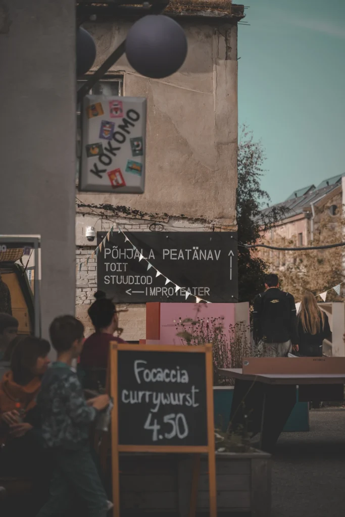 A corner of the Pohjala Factory courtyard with direction signs, a Kokomo sign, a menu board and people sitting outside.