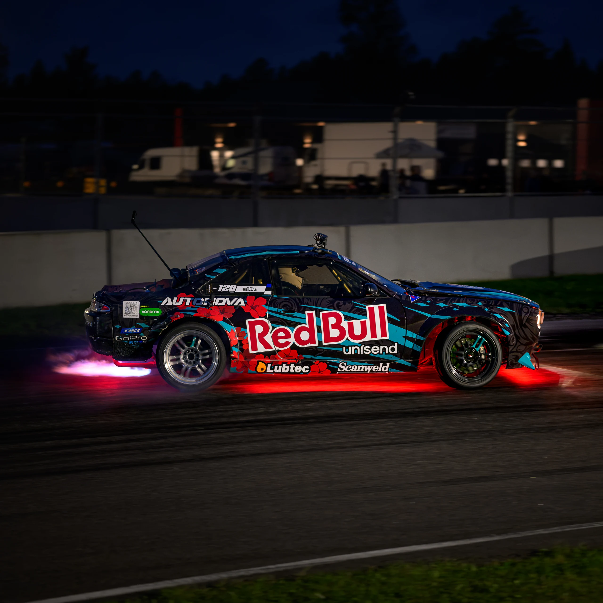 A drift car with colourful decals on the track at night, with a red glow underneath and a pink flame behind it.