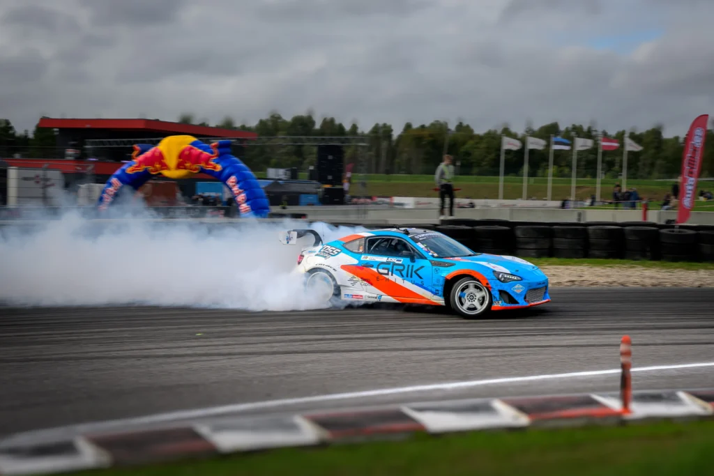 Sinioranž driftiauto veab üle raja suitsujuti, taustal lipud ja täispuhutav võistluskaar.