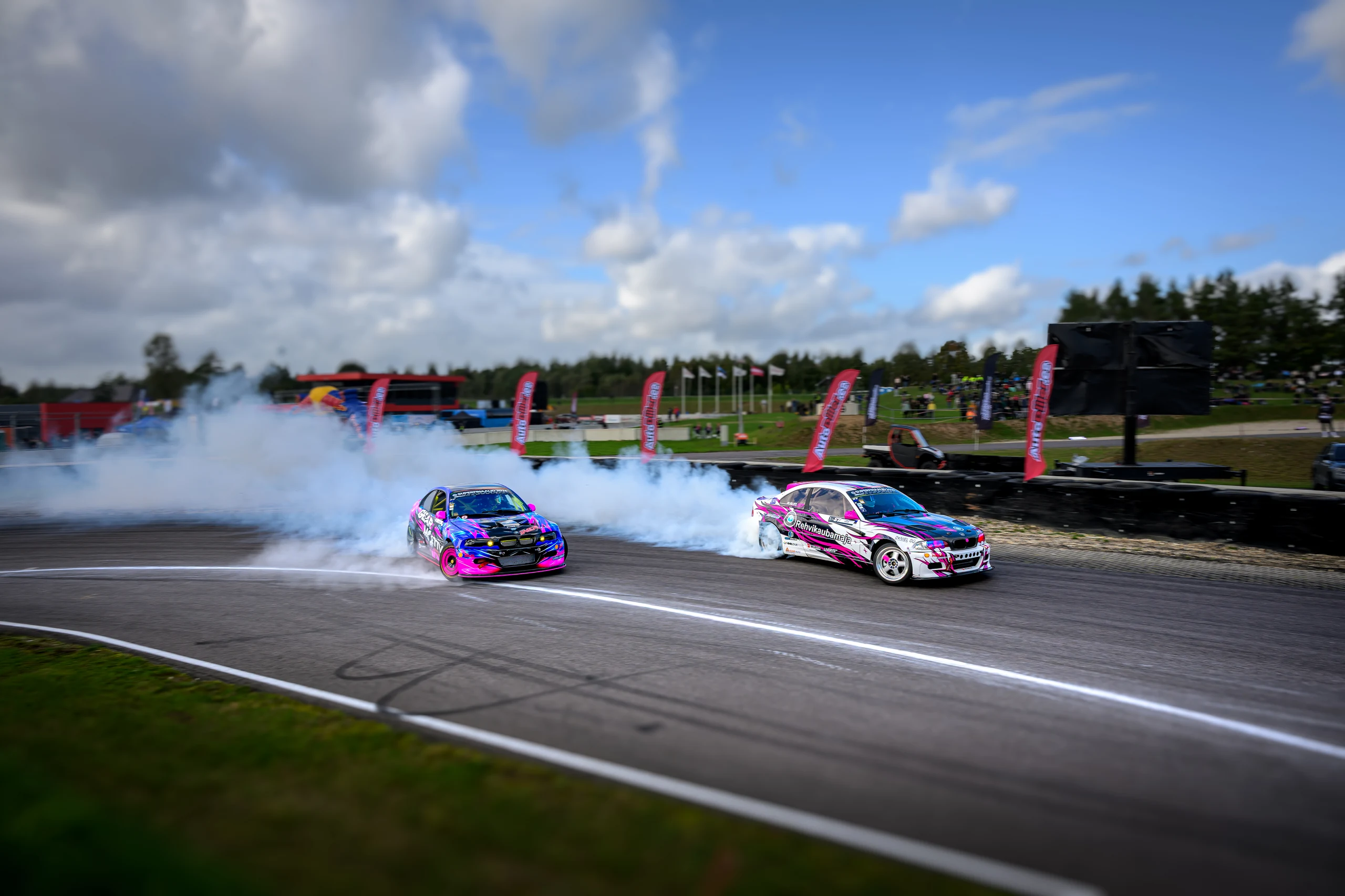 Two drift cars with pink, blue and white liveries taking a corner at Raassilla in daylight, trailing a broad cloud of smoke.
