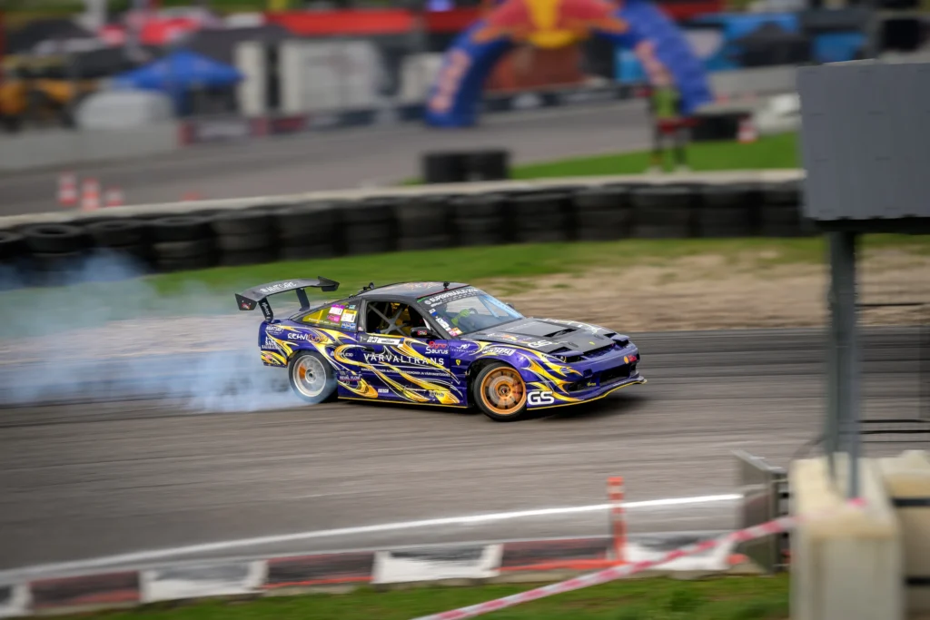 Suure tagatiivaga lillakas driftiauto liigub üle raja, selle kuldsed veljed ja taust on liikumisest hägusad.