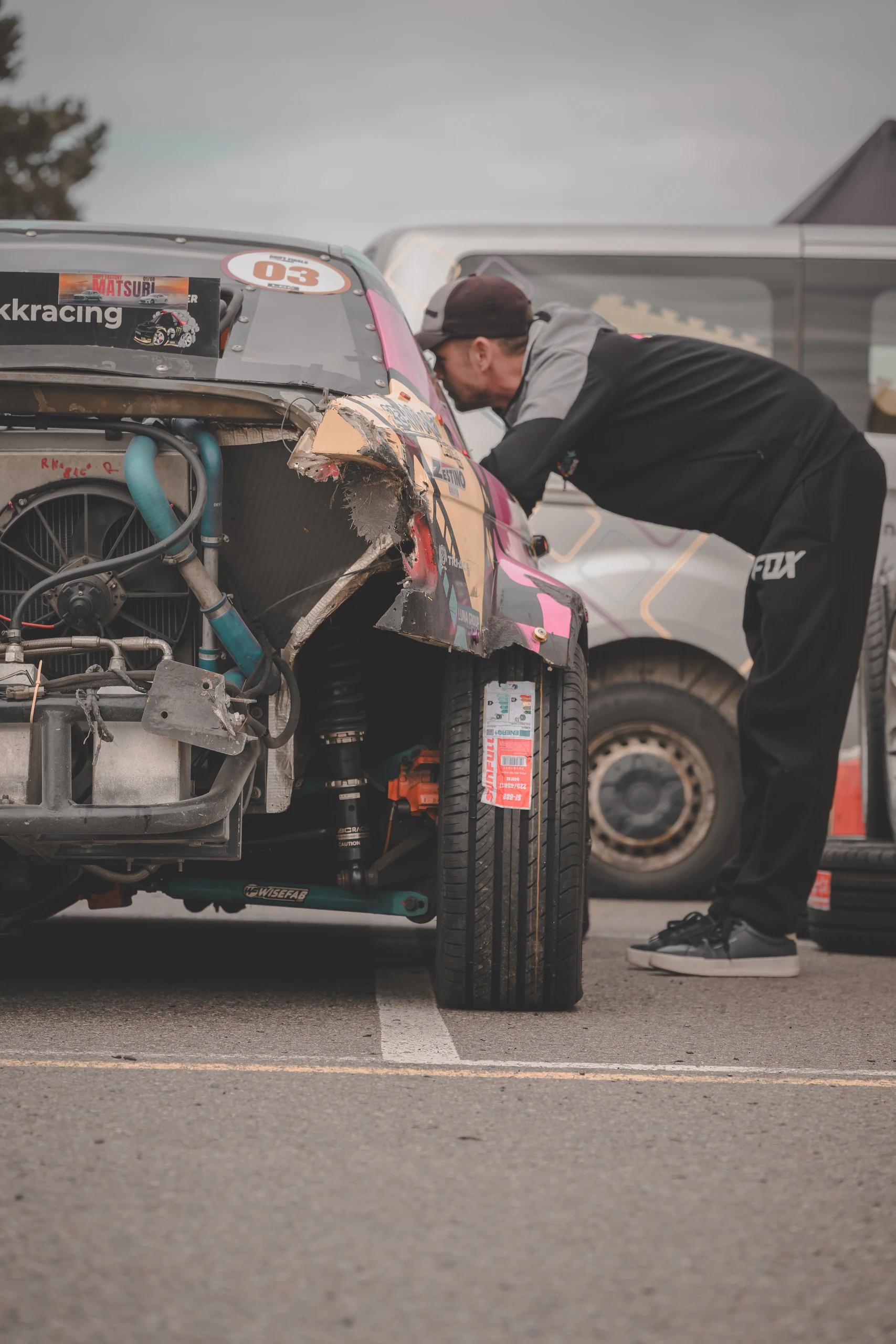 A man in a cap leans towards a drift car, with a fan and blue hoses visible in an exposed section of the car.