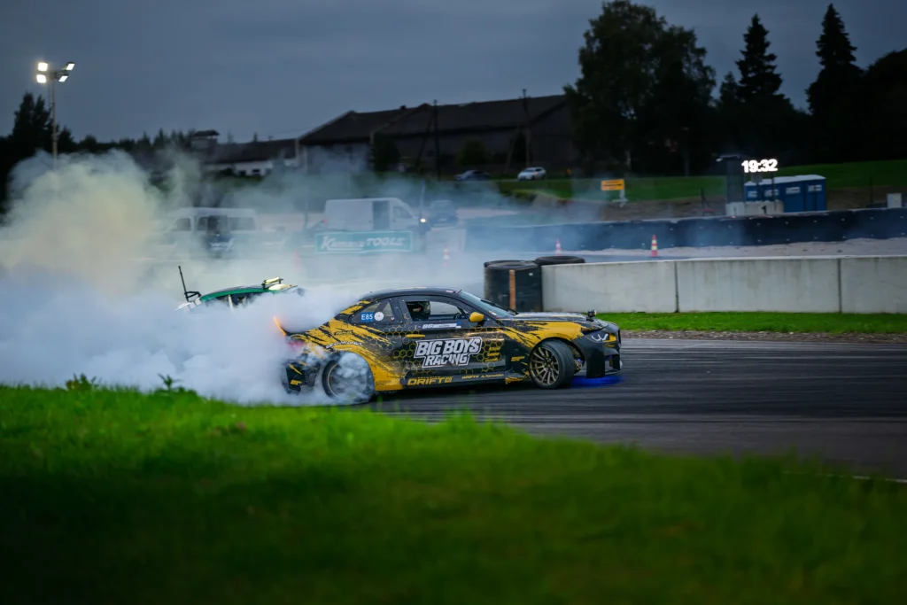 Mustkollane driftiauto ning osaliselt suitsu peitunud roheline auto hämaral rajal.