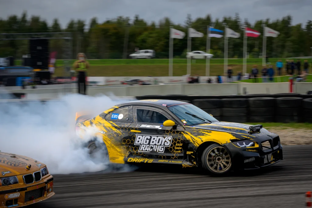 Mustkollane driftiauto möödub rehvidest rajapiirdest, lipud ja puud taustal.