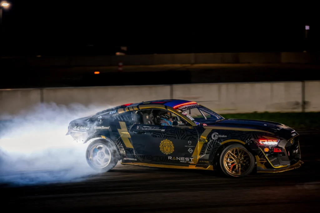 Mustkuldne driftiauto valgustatud suitsu ees pimedal rajal, kuldsed veljed eristumas külgvaates.