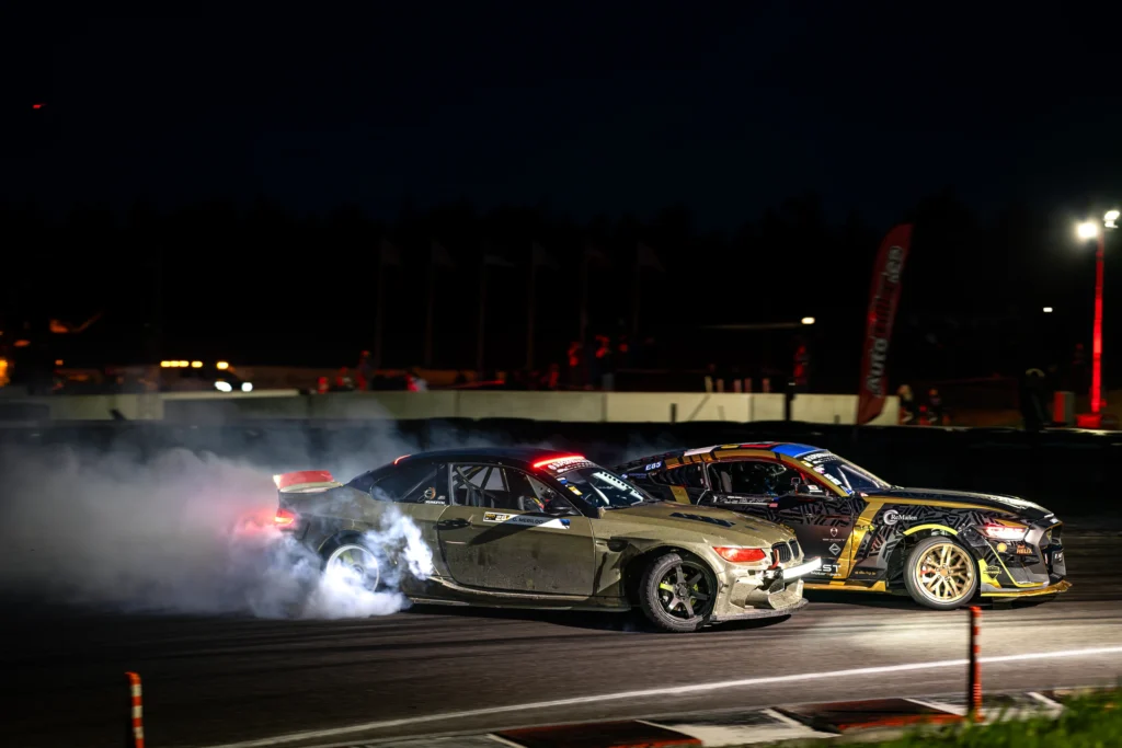 Oliivroheline BMW ja mustkuldne driftiauto libisevad öösel kõrvuti, valge suits ja punased tagatuled nende taga.