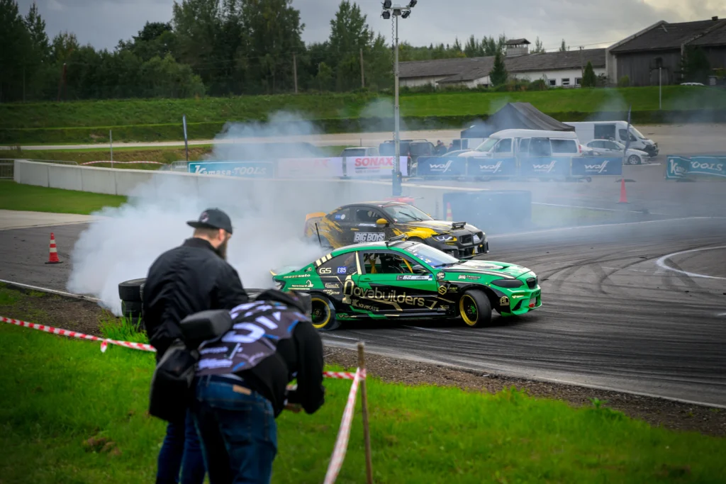 Kaks pealtvaatajat jälgivad rajapiirde äärest rohelist ja mustkollast driftiautot kurvis.