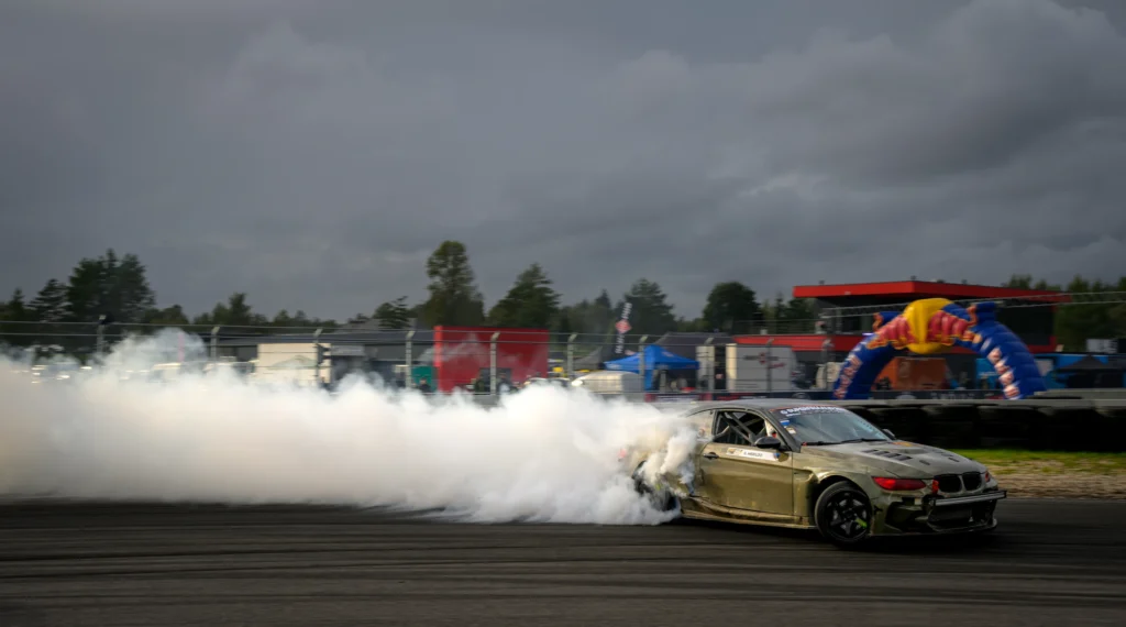 Oliivroheline driftiauto pika valge suitsujuti ees, rajal taustaks võistluskaar ja lipud.