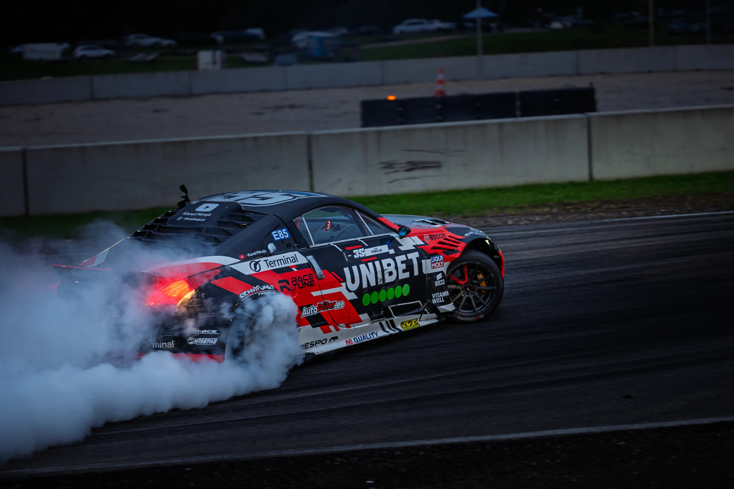 A red, black and white drift car on the Raassilla track, with thick tyre smoke around its rear wheels.