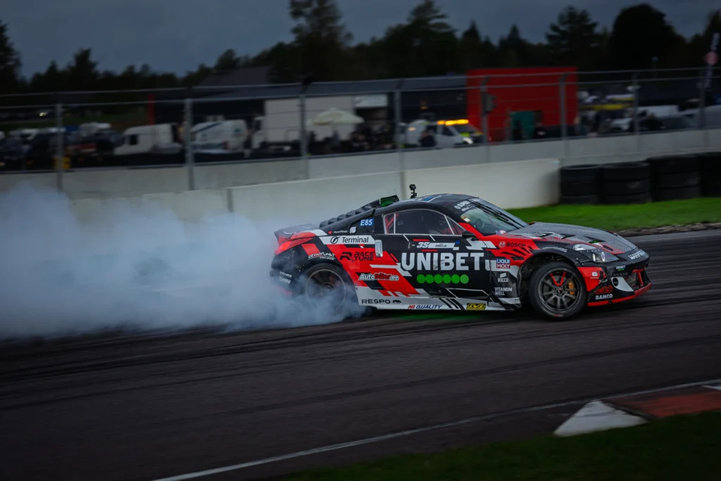 Punase ja musta kujundusega driftiauto suitsujuti ees hämarduval rajal.