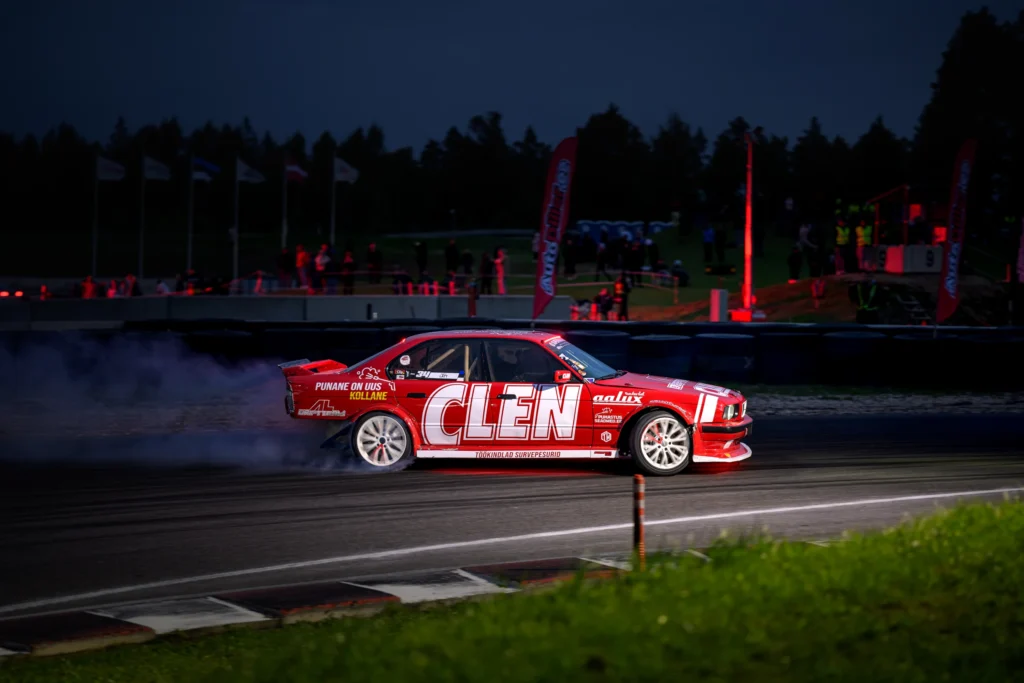 Punane valgete velgede ja suure CLEN-kirjaga driftiauto valgustatud rajal.