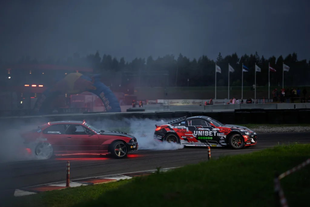 Punane auto järgneb punamustale driftiautole sinakashallis suitsus.