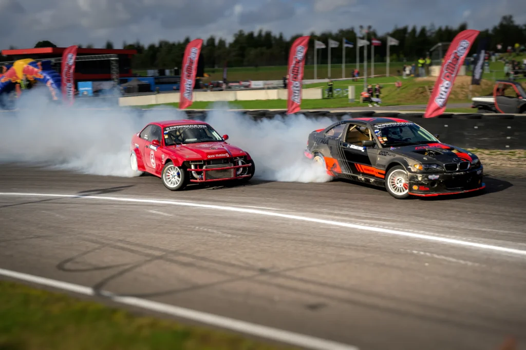 Must-oranž driftiauto liigub punase auto ees; asfaldil on tumedad rehvijäljed ja taustal punased lipud.