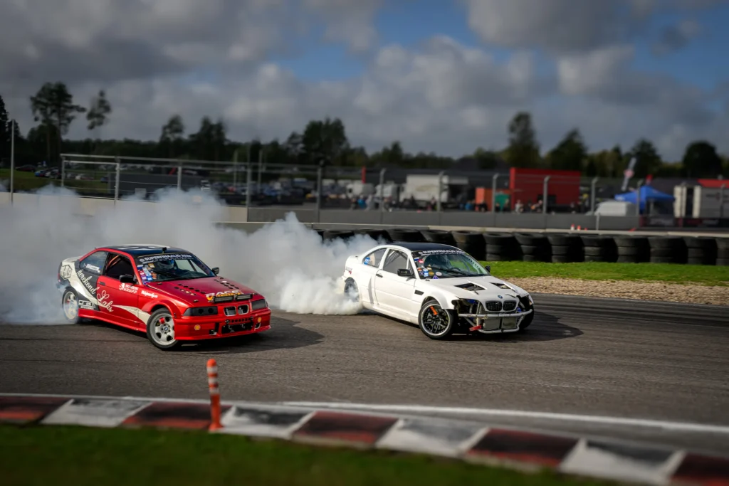 Punane ja valge BMW sõidavad kõrvuti läbi kurvi, nende taga lai rehvisuitsupilv ja pilvine taevas.
