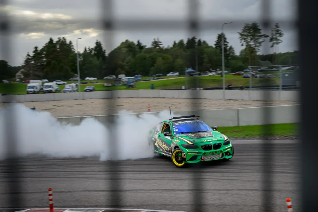 Roheline driftiauto läheneb läbi laia valge suitsupilve, taustal rajahoone ja puud.