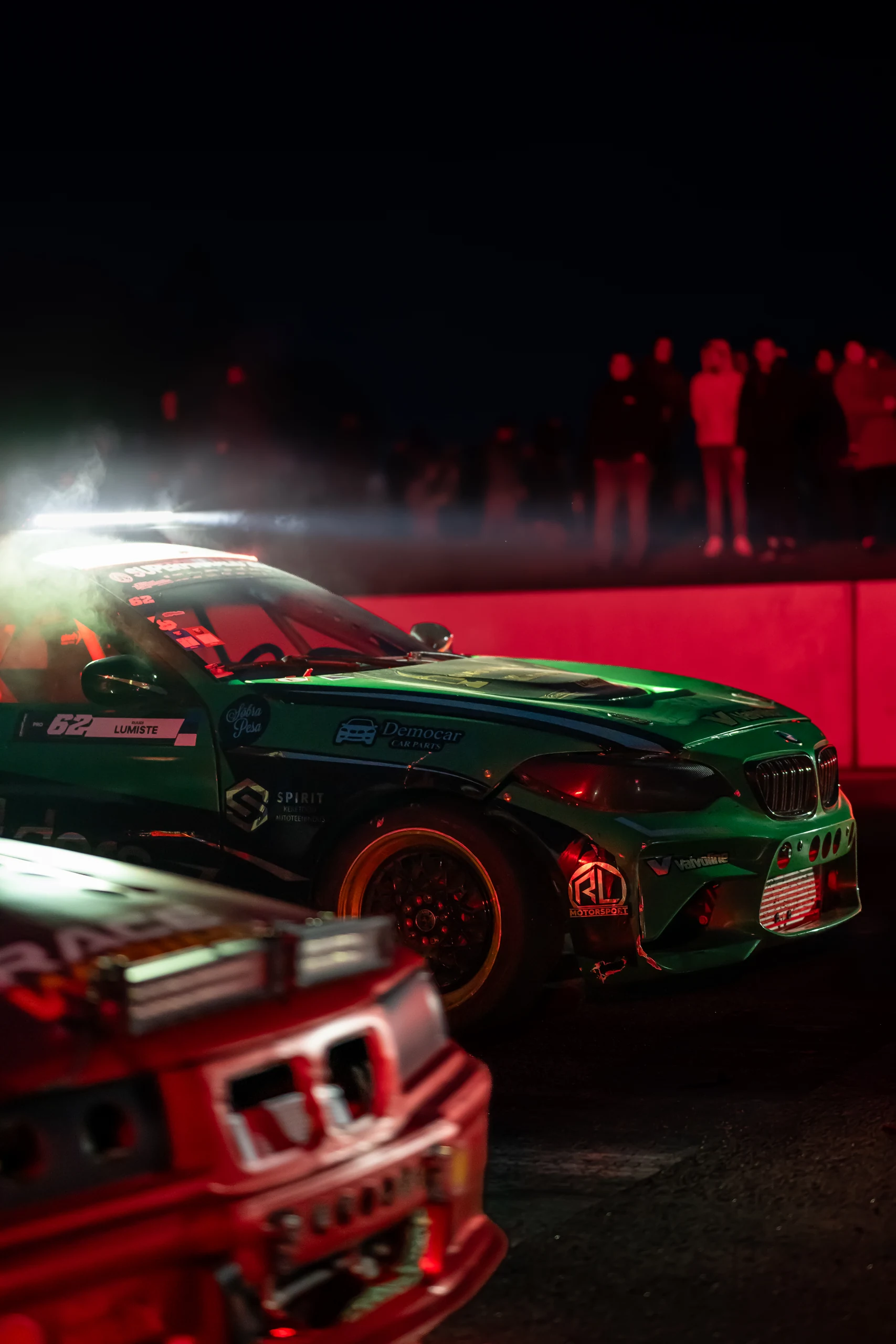 A green drift car in red light at night, with smoke behind it, part of another car in the foreground and spectators beyond the track.
