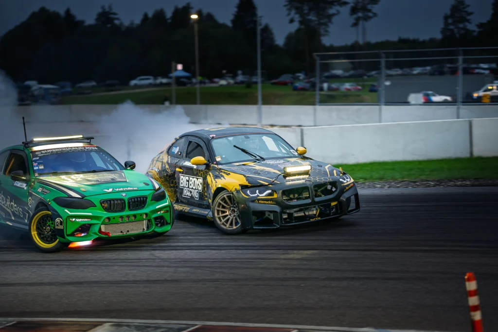 Roheline ja mustkollane driftiauto liiguvad lähestikku külg ees, suitsupilv nende taga.