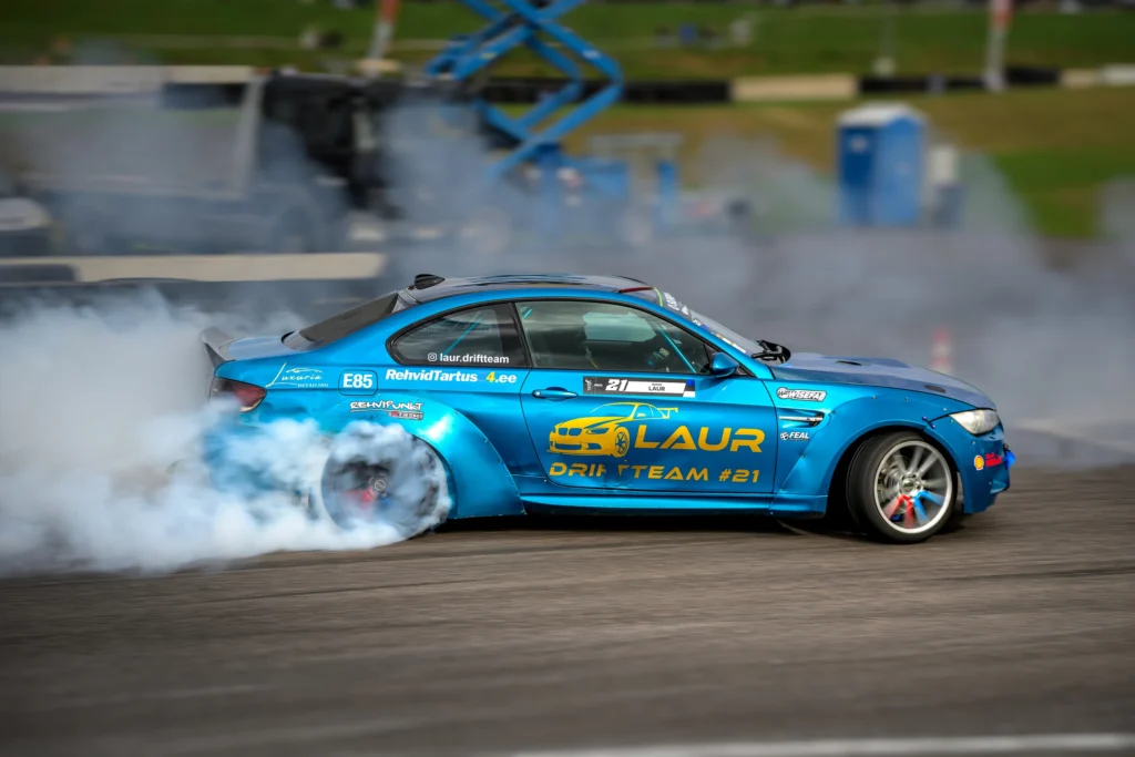 Ere sinine BMW kollase küljekirjaga driftib külg ees, tagaratta juurest tõuseb valget suitsu.