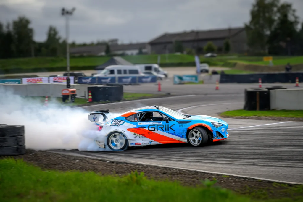 Sinise, valge ja oranži kujundusega driftiauto libiseb külg ees, tagarataste taga valge suits.