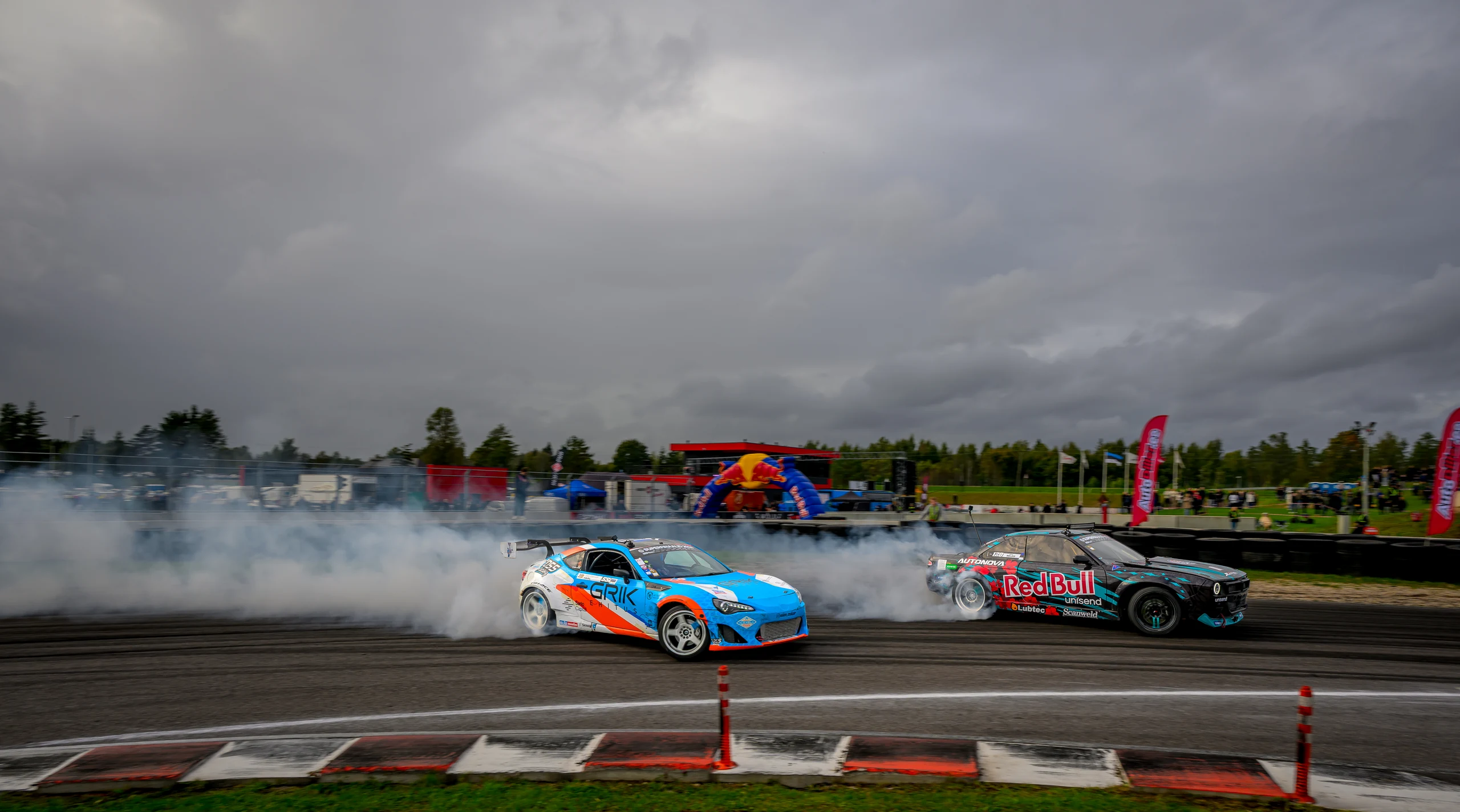 Sinioranž ja tume driftiauto läbivad koos kurvi, suitsuriba tumeda pilvetaeva all.
