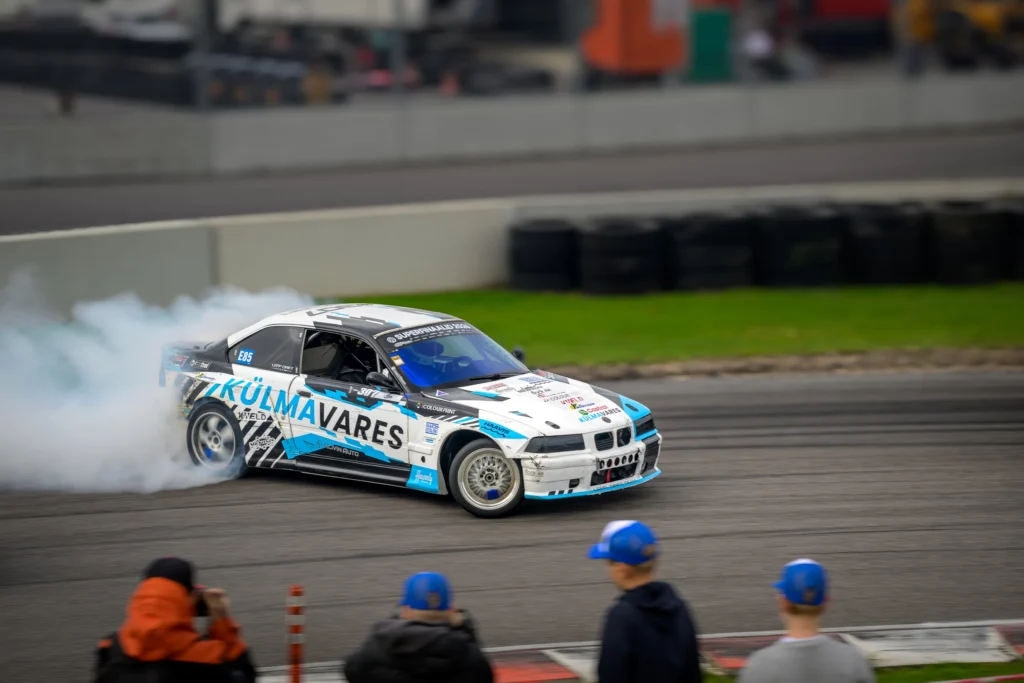 Valge-sinine auto libiseb suitsupilve ees; kaadri allservas jälgivad sõitu pealtvaatajad.
