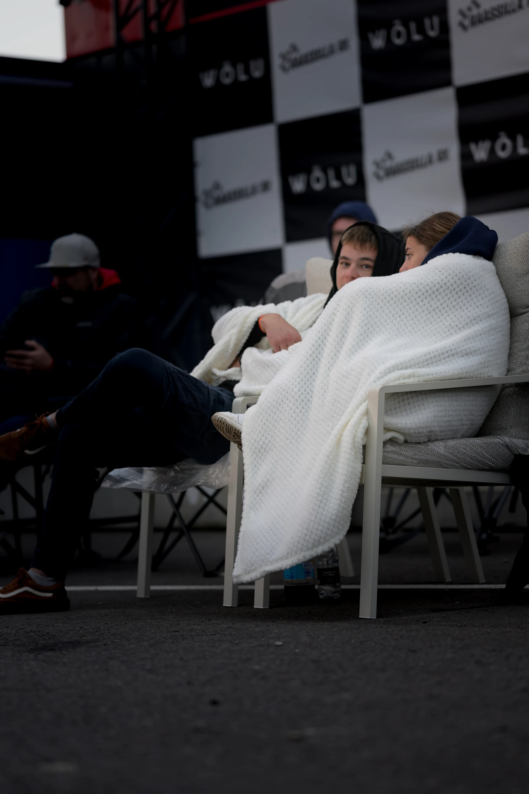 Two people sit side by side in a paddock tent wrapped in white blankets, with other people in the background.