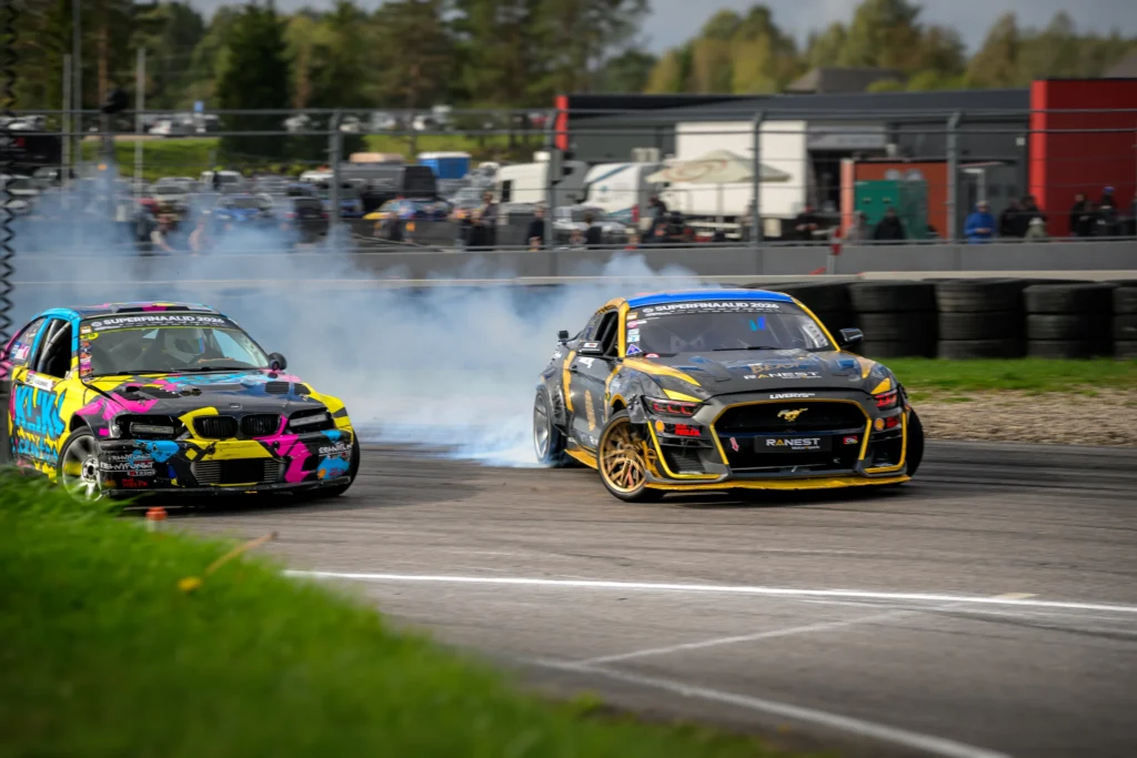 Kirju kujundusega auto ja must kollaste ääristega auto driftivad lähestikku üle rajajoonte.
