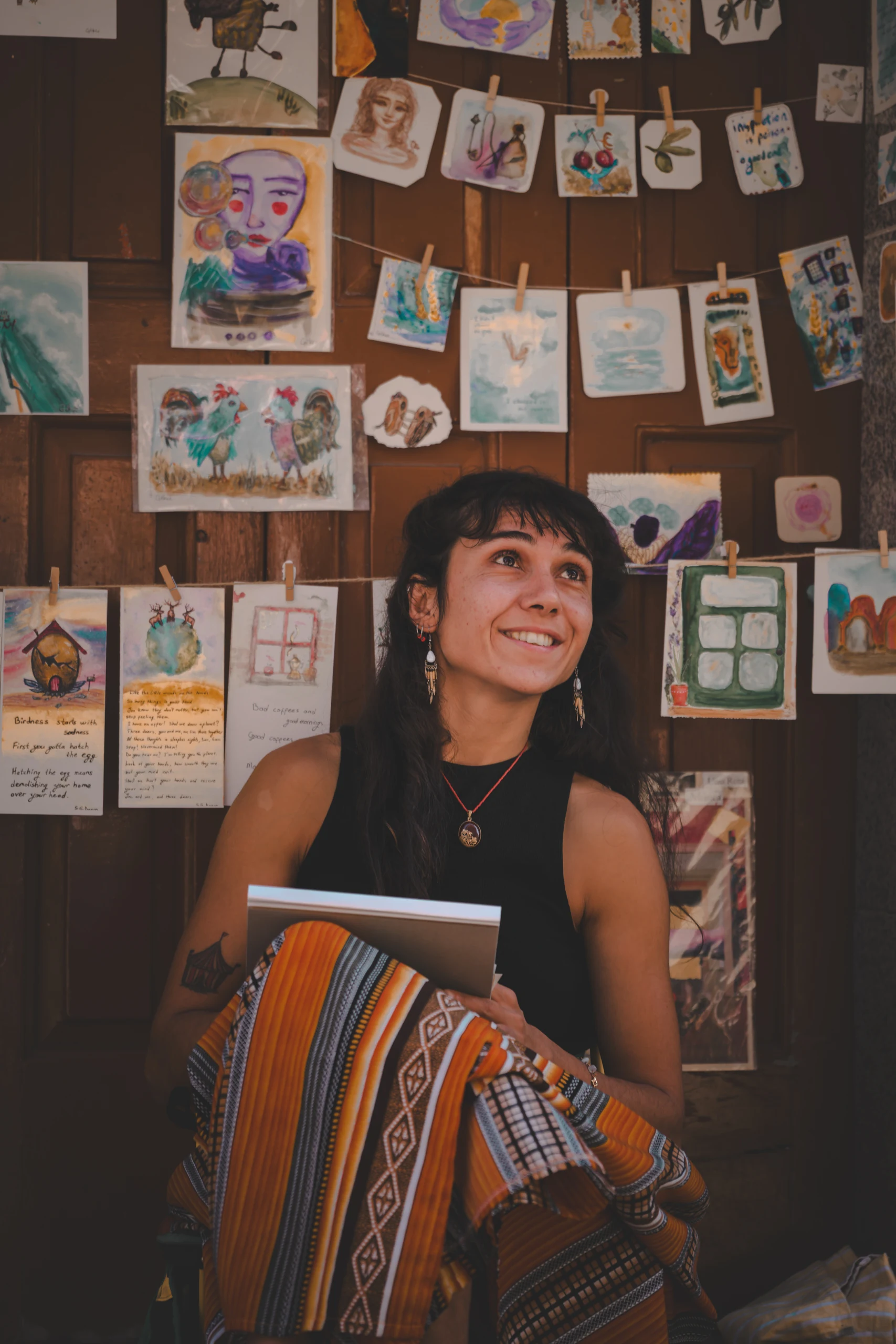 Gökçe sits in front of a wooden door covered in colourful pictures, with striped fabric and a sketchpad on her lap.