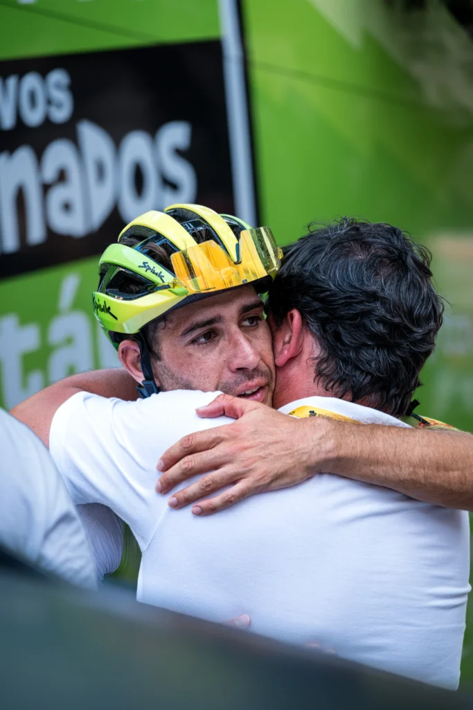 A cyclist in a yellow helmet embraces a man with a hand on his shoulder.