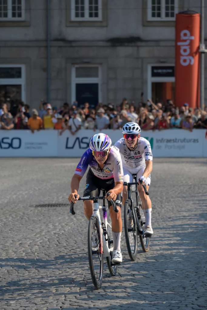 Two cyclists in white helmets ride one behind the other over the cobbles.