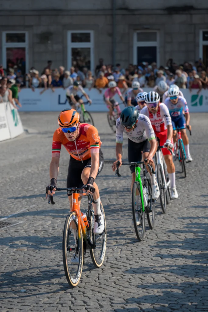 A cyclist in an orange jersey leads a small group out of a cobbled bend.