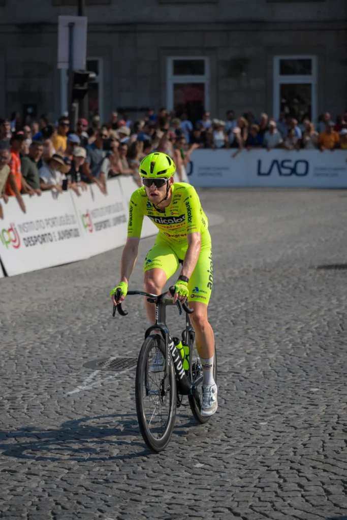 A cyclist in fluorescent yellow racing kit approaches with spectators and a stone facade behind.