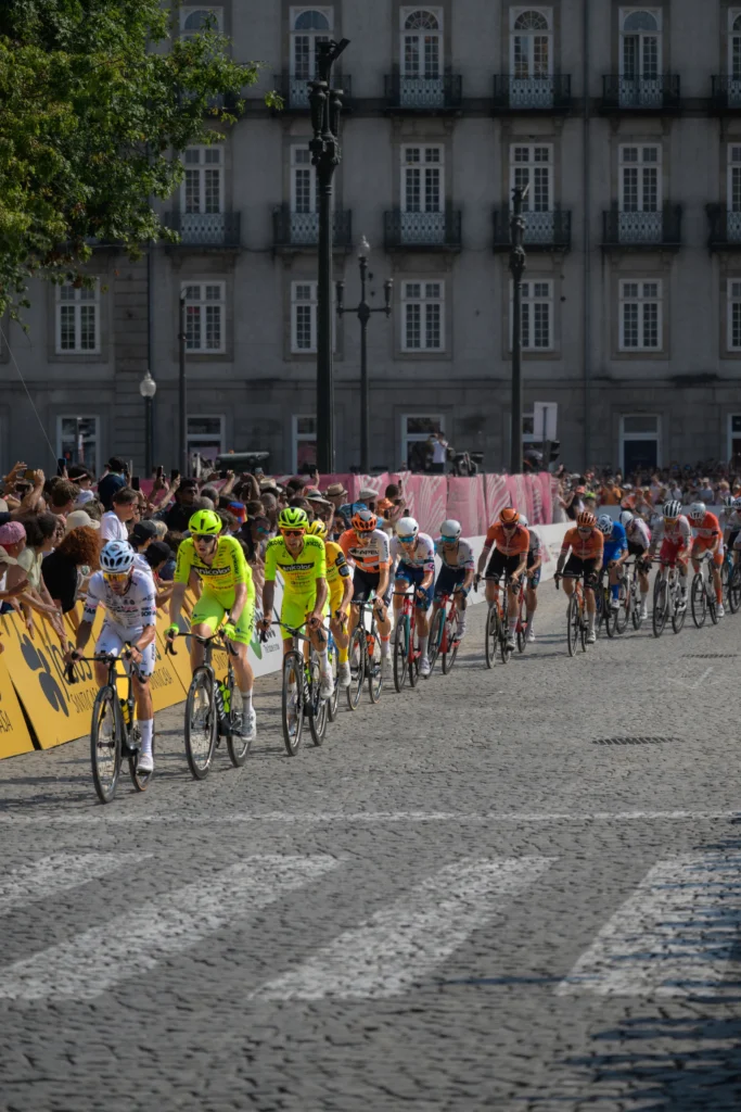 A line of cyclists in bright racing jerseys rounds a bend in front of spectators.