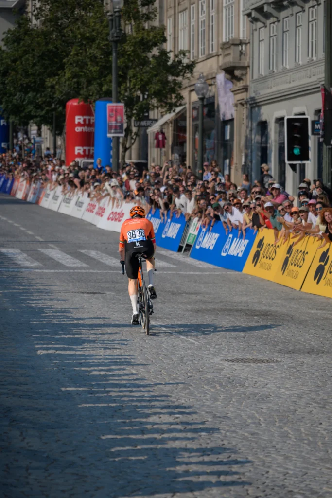 A lone cyclist in an orange jersey rides away along a straight lined with spectators.