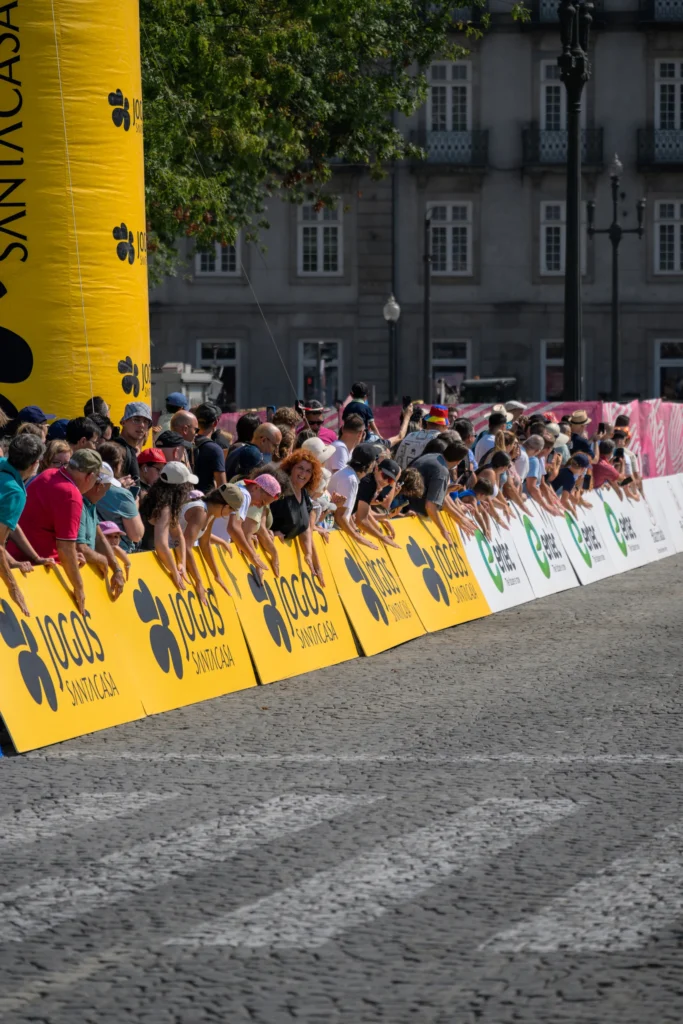 Spectators watch the race from behind yellow barriers beside the cobbled road.