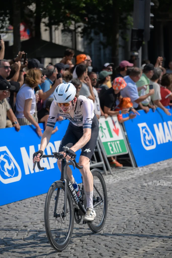 A cyclist in a white helmet passes spectators standing behind blue advertising barriers.