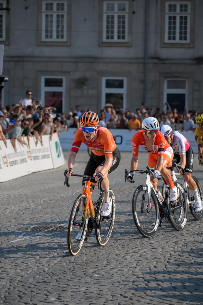Cyclists in orange and red racing kit ride side by side around a bend in front of spectators.
