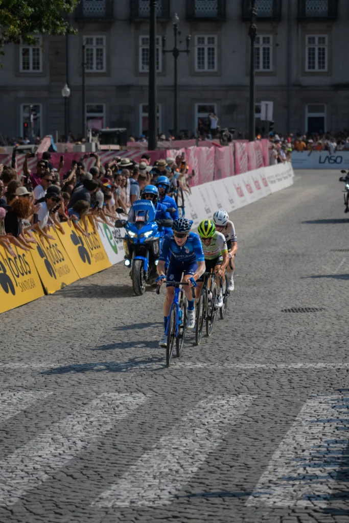 Cyclists and a blue escort motorcycle approach along a street lined with spectators.