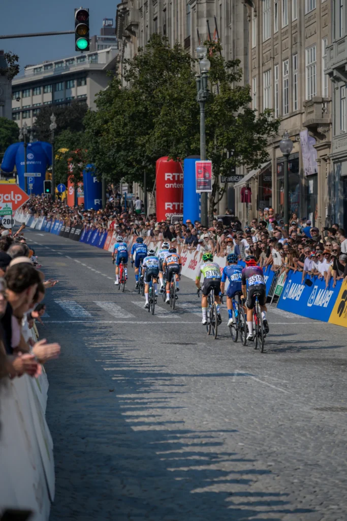A long line of cyclists follows a street through crowds of spectators in Porto.
