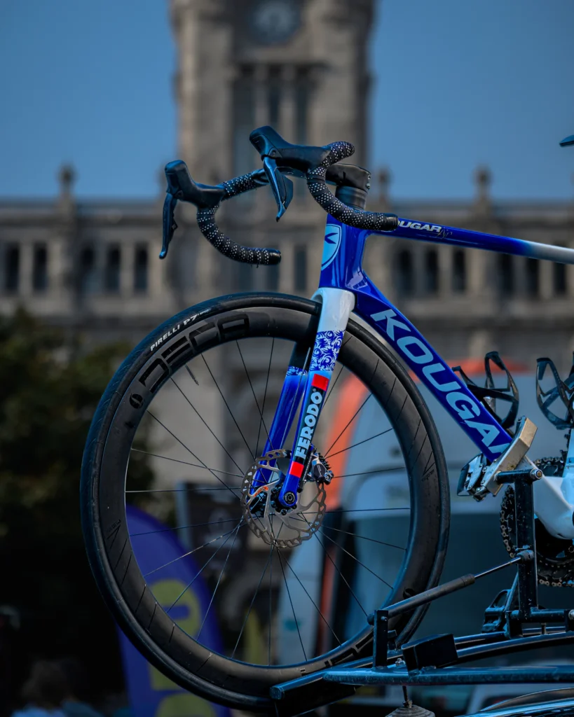 The front wheel and handlebars of a blue road bicycle with Porto city hall blurred behind.