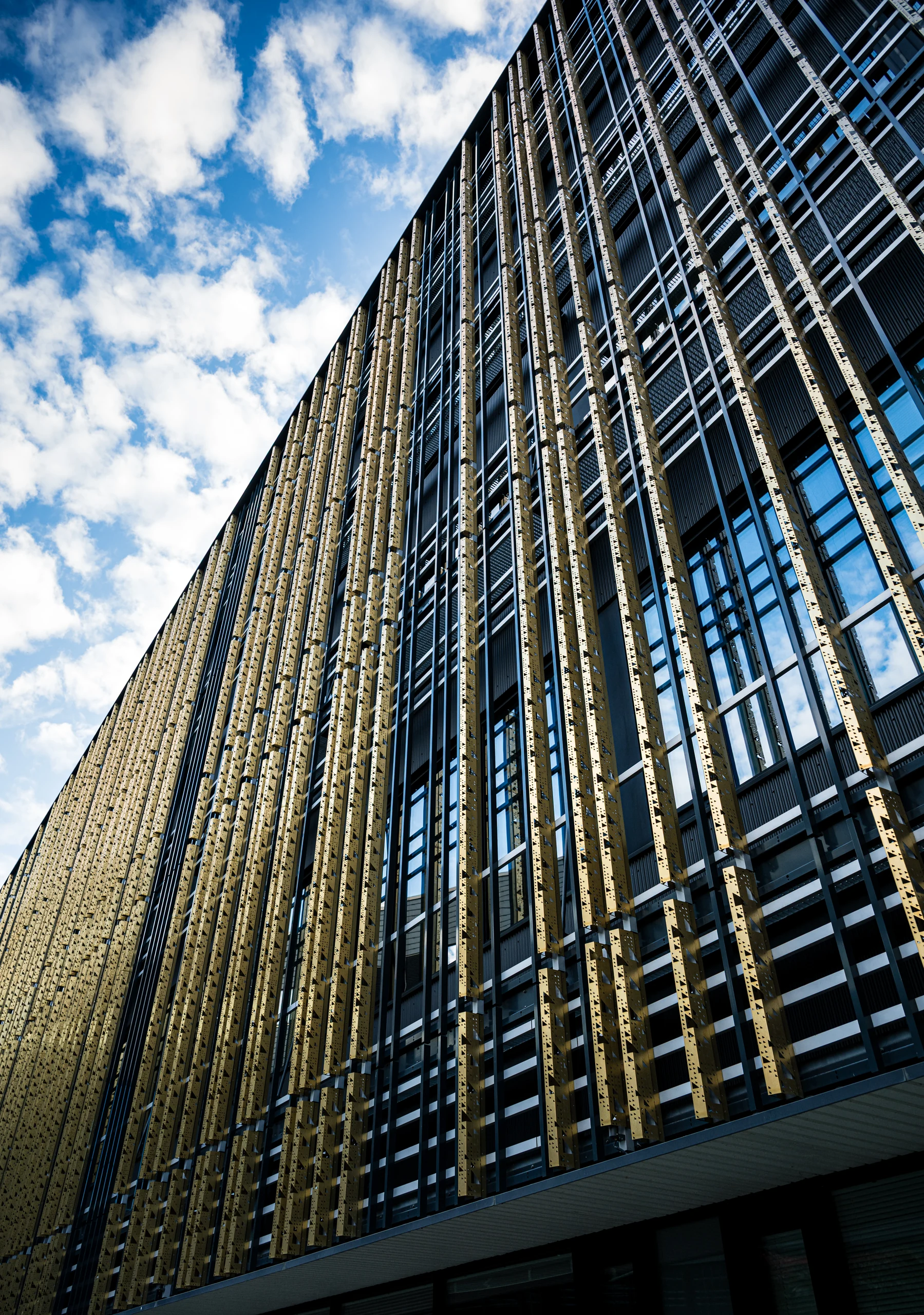 A diagonal view up the Delta Centre’s tall facade with golden fins, beside blue sky and white clouds.